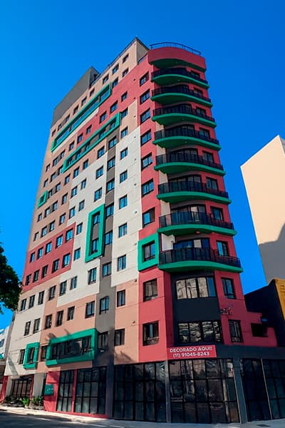Fachada do edifício com arquitetura colorida em tons de vermelho, verde e cinza, sob céu azul.